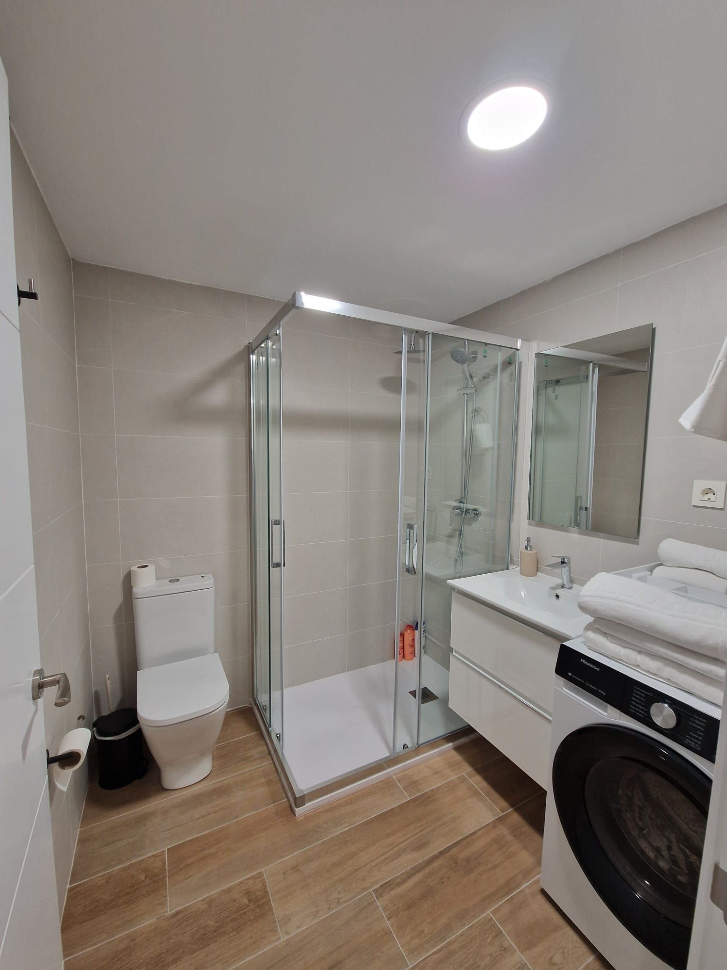 Baño moderno y luminoso con ducha de cristal, inodoro, lavabo y lavadora-secadora apilada con suelo de imitación madera.