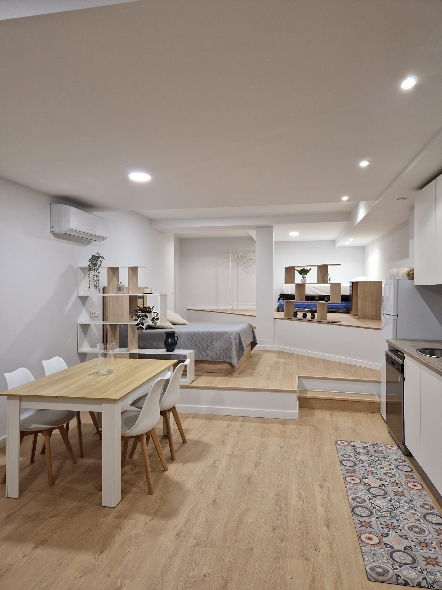 Apartamento tipo estudio luminoso y diáfano con mesa de comedor, cama sobre plataforma, sofá y suelos de madera.