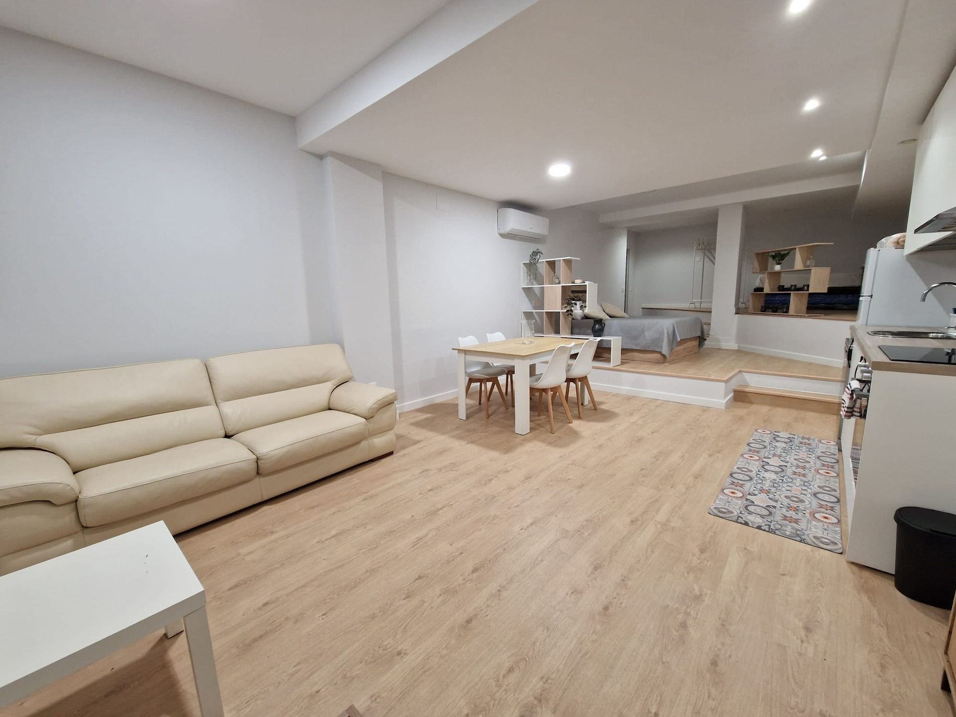 Sala de estar luminosa y abierta con sofá seccional beige, pisos de madera, mesa de comedor y cocina al fondo.