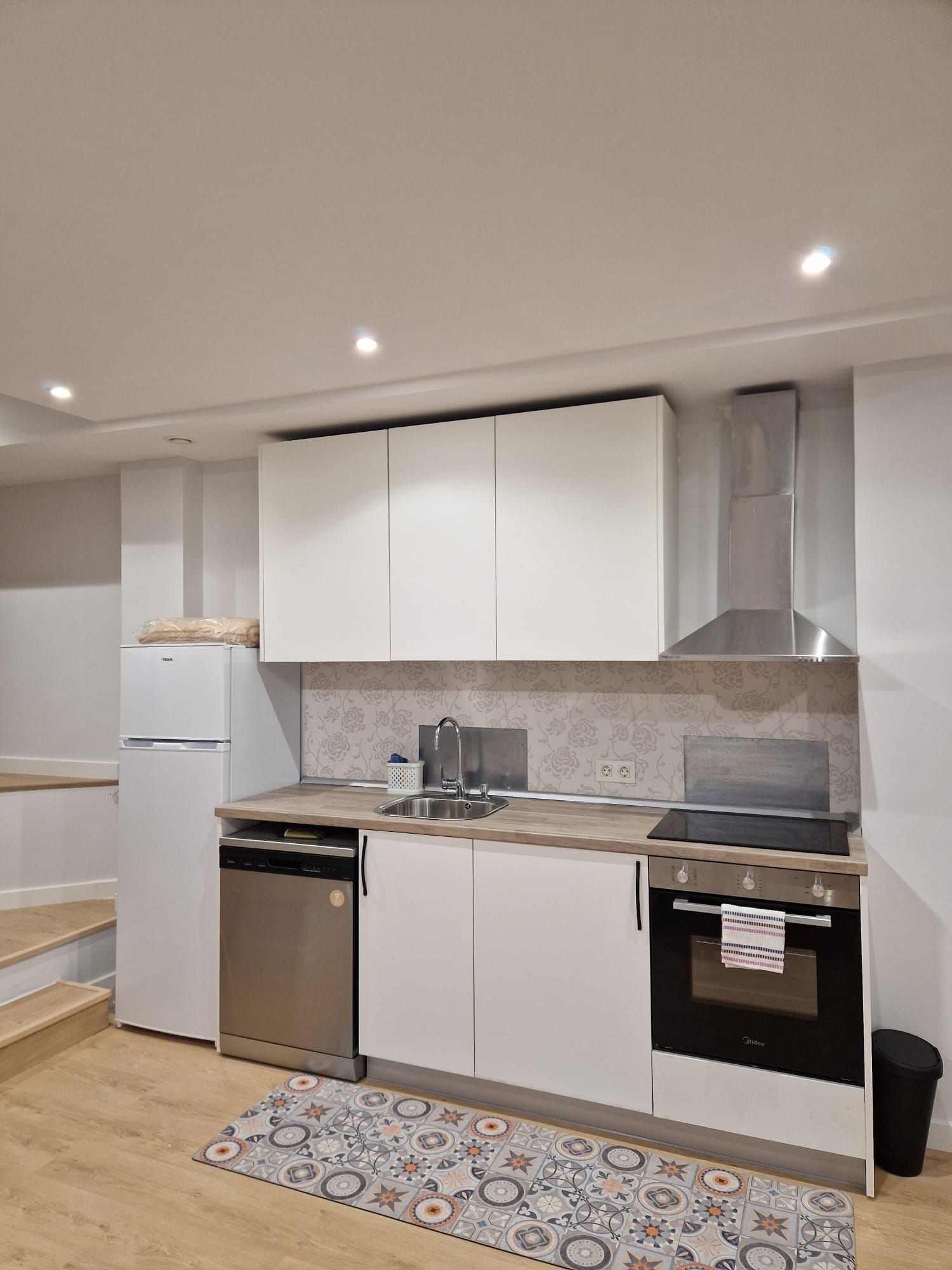 Cocina moderna con armarios blancos, electrodomésticos de acero inoxidable, suelo de madera y una pequeña alfombra estampada.