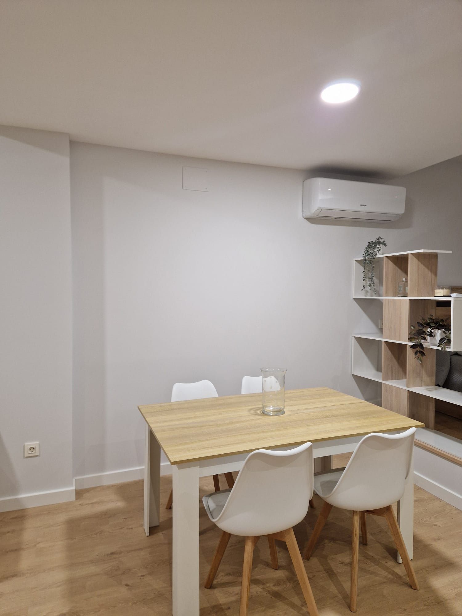 Pequeño comedor con mesa de madera, sillas blancas, estante de pared y aire acondicionado en una habitación luminosa.