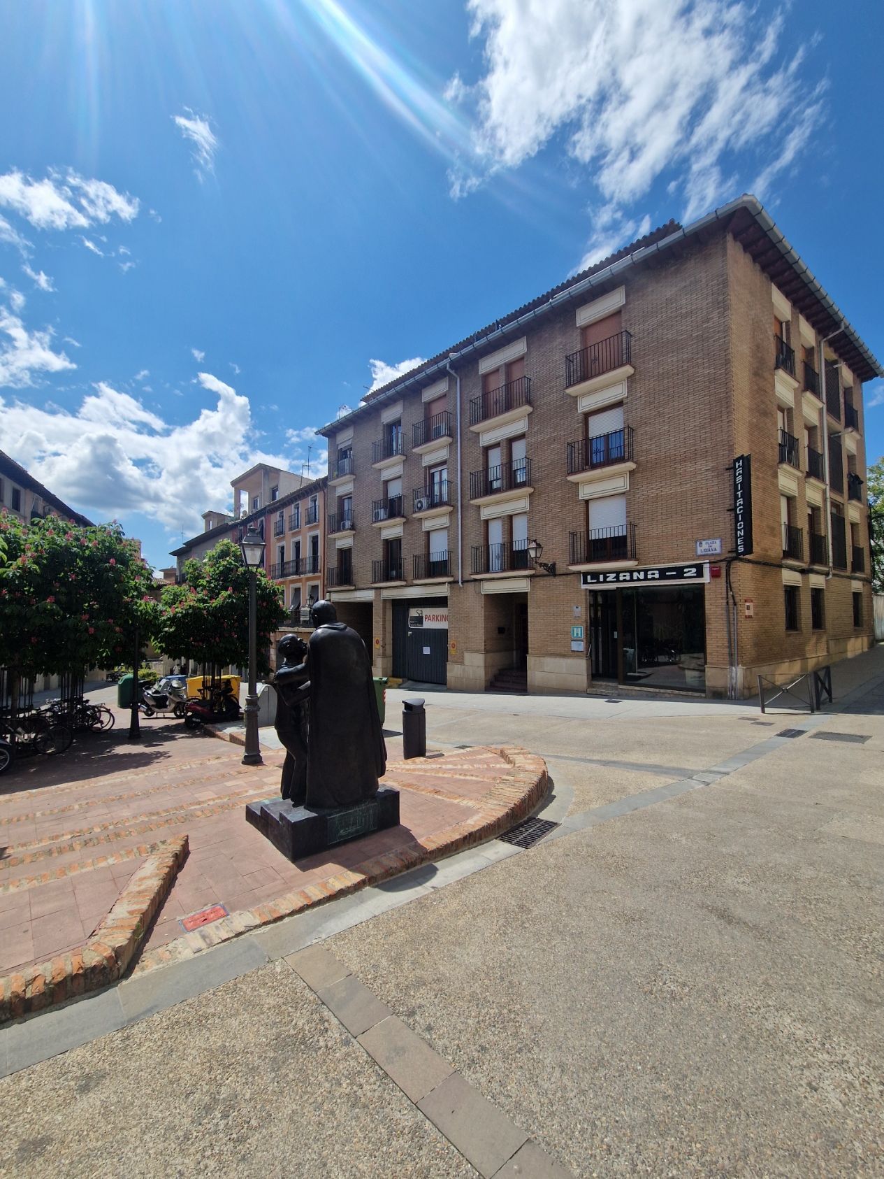 Edificio histórico de ladrillo en una esquina soleada con una pequeña fuente o escultura en primer plano.