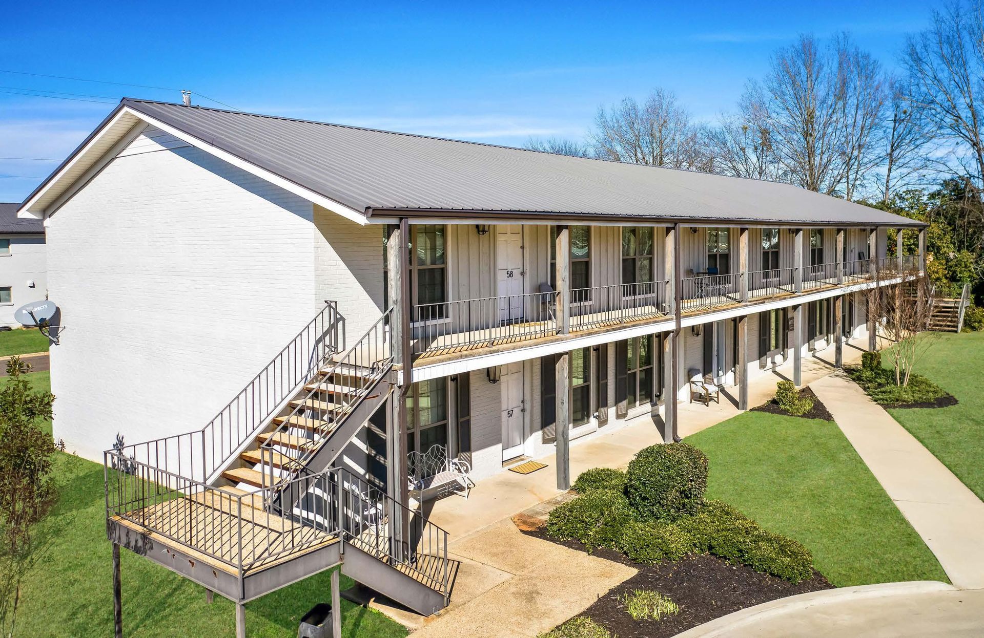 Two-story apartment building with exterior walkways, stairs, and landscaped grounds under a clear blue sky