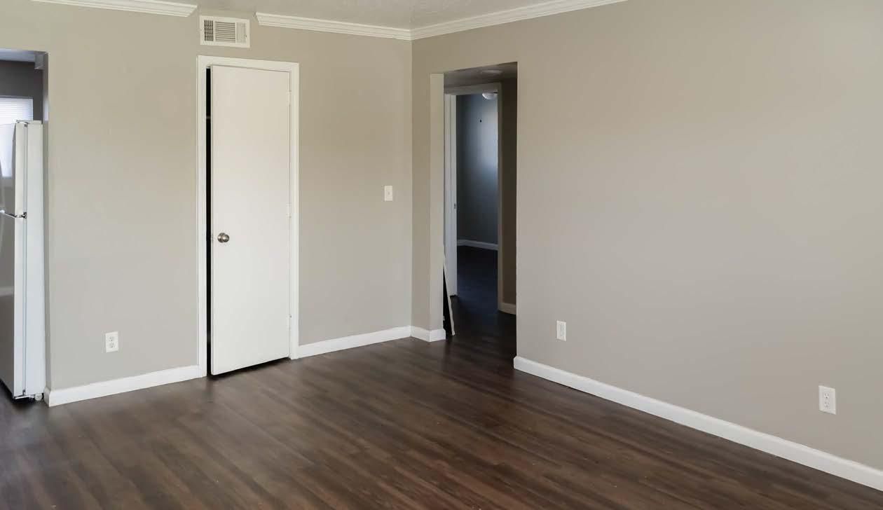 Empty living room with dark wood floors, beige walls, white doors, and a view into a hallway.