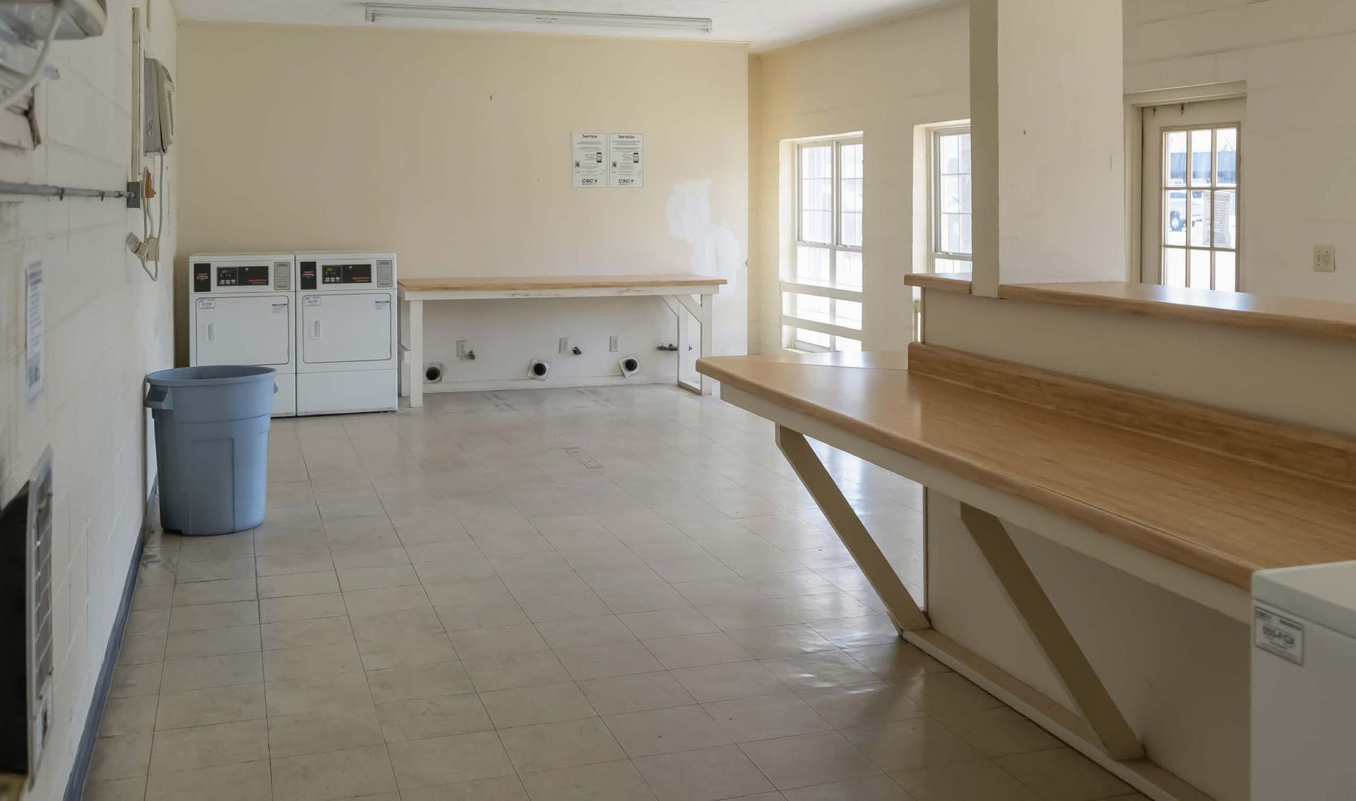 Empty utility room with white tile floor, beige walls, worktables, washers, and a trash can near windows.