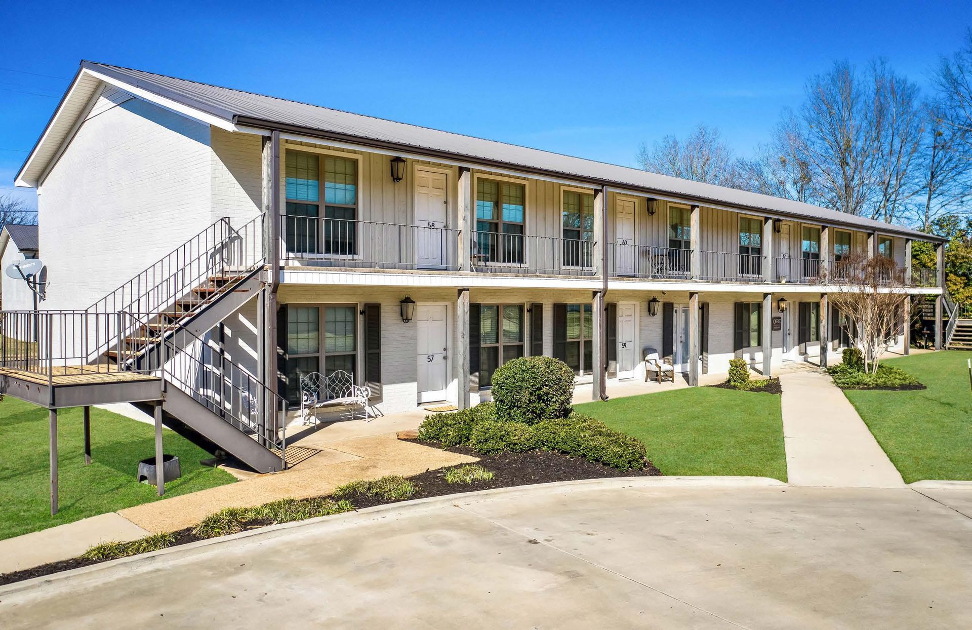 Two-story motel with exterior corridors, stairs, and landscaped grounds under a clear blue sky