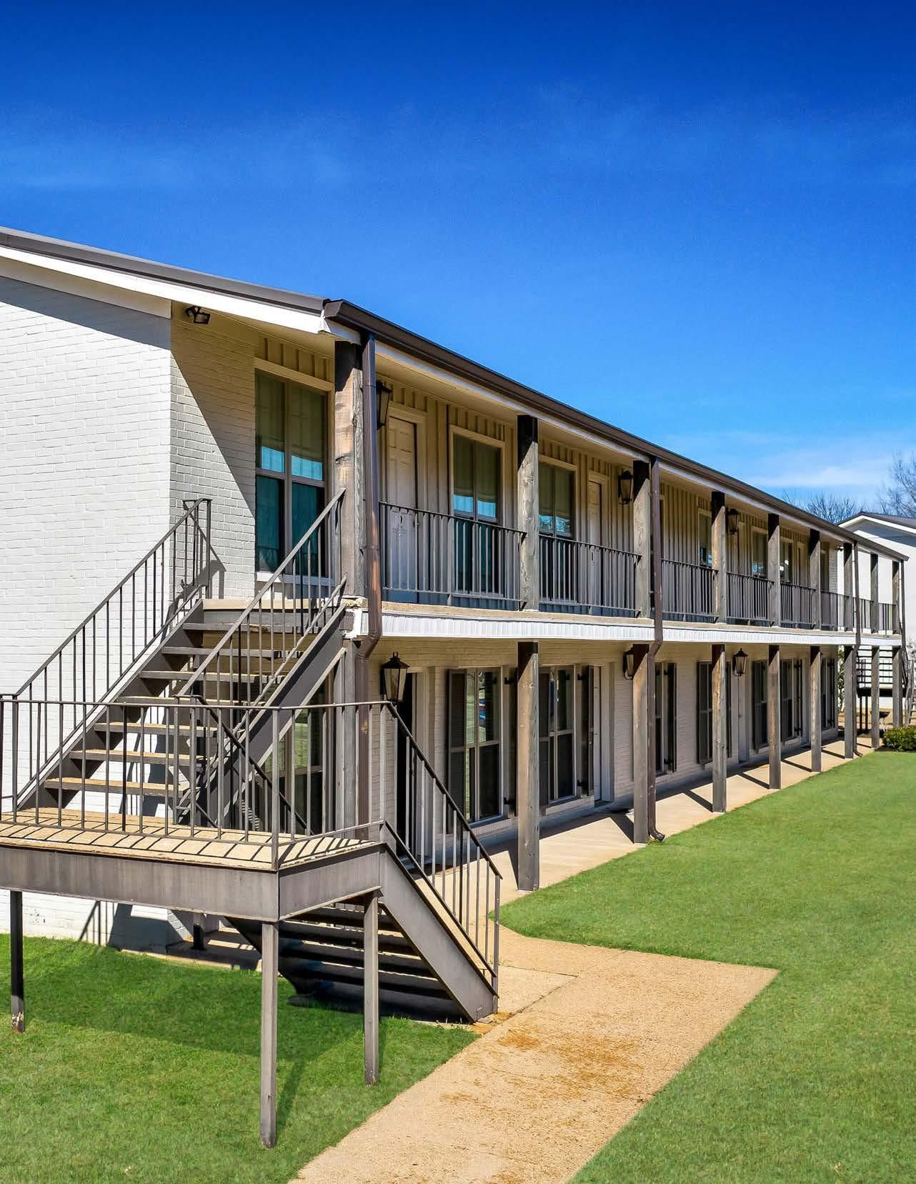 Two-story motel-style building with exterior stairways and balconies beside a grassy walkway under a blue sky