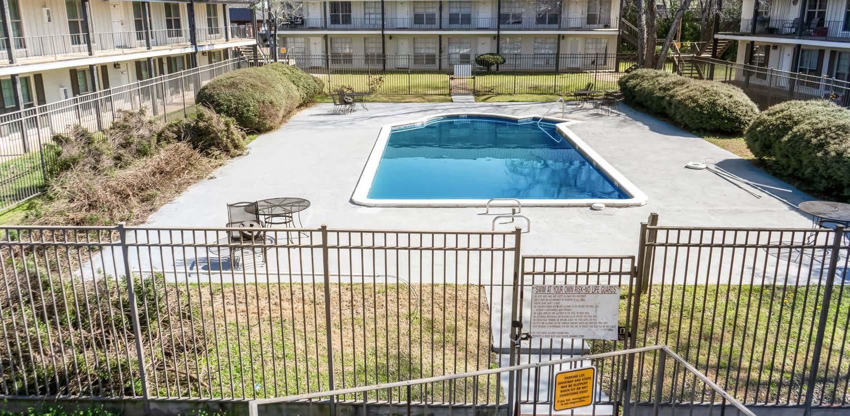 Apartment pool surrounded by a fence and patio seating, with a building in the background