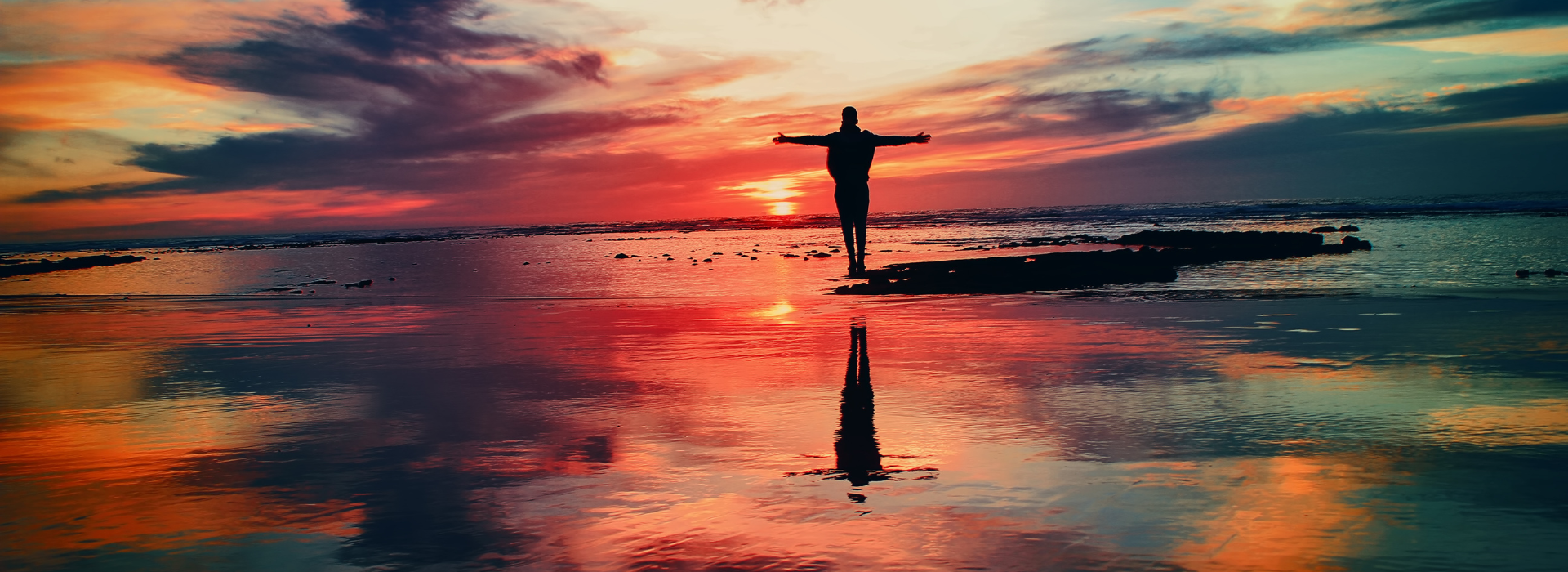 A person is standing on a beach at sunset with their arms outstretched.