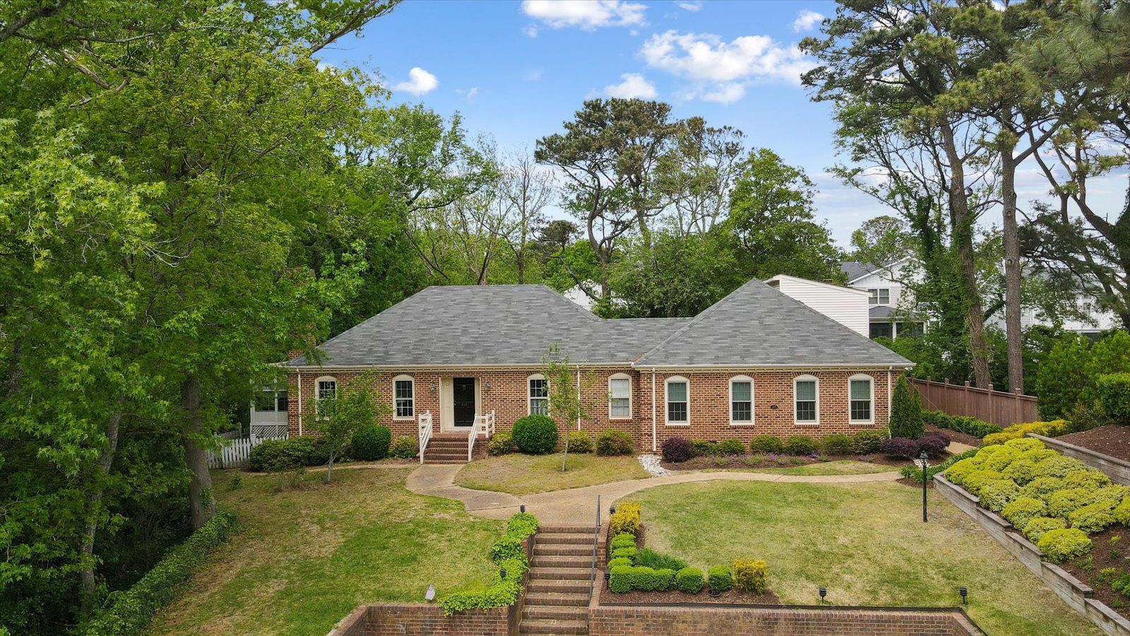 Brick house with gray roof, front steps, and landscaped yard surrounded by trees under a blue sky