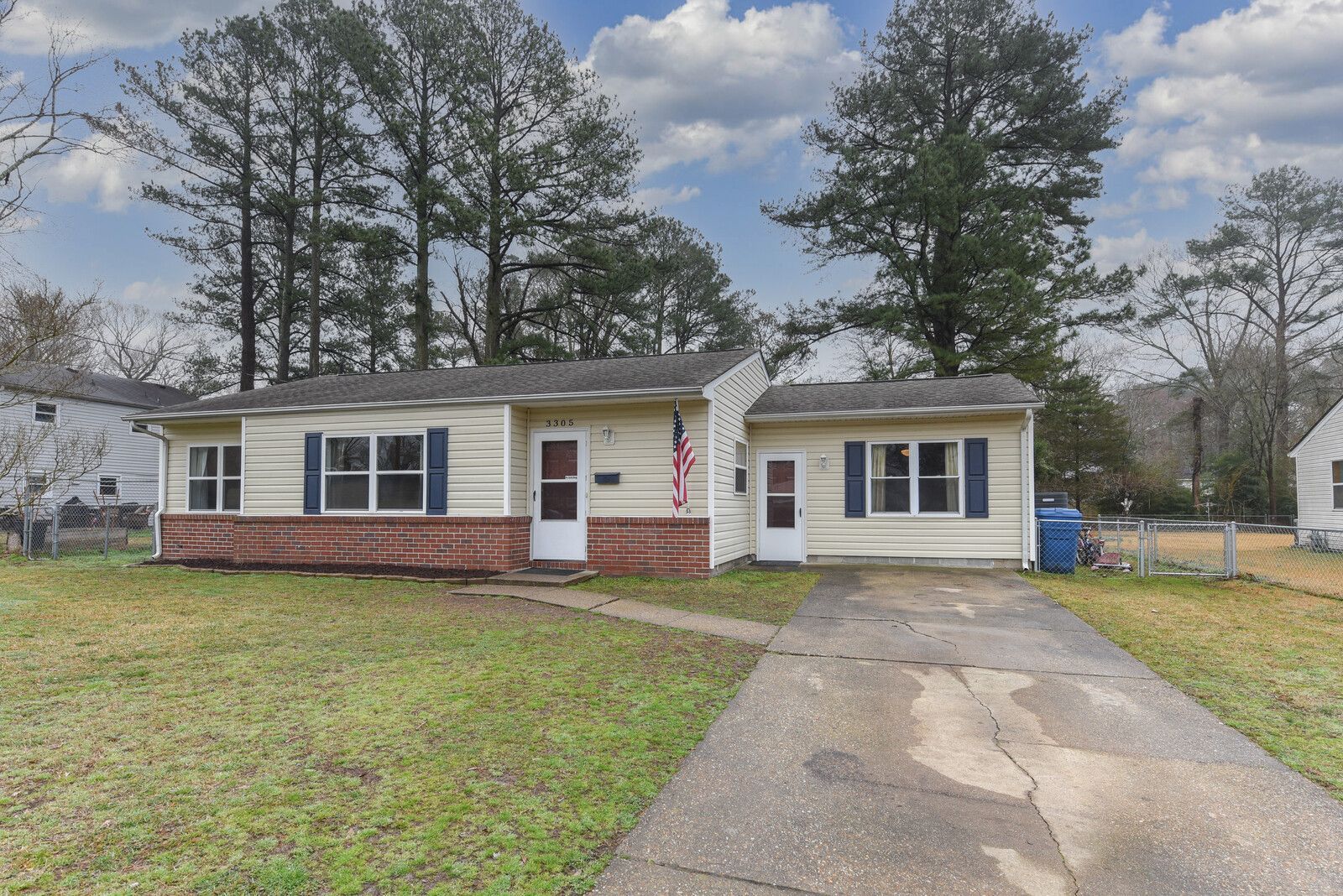 Single-story suburban house with white siding, red brick, and a driveway under cloudy skies.