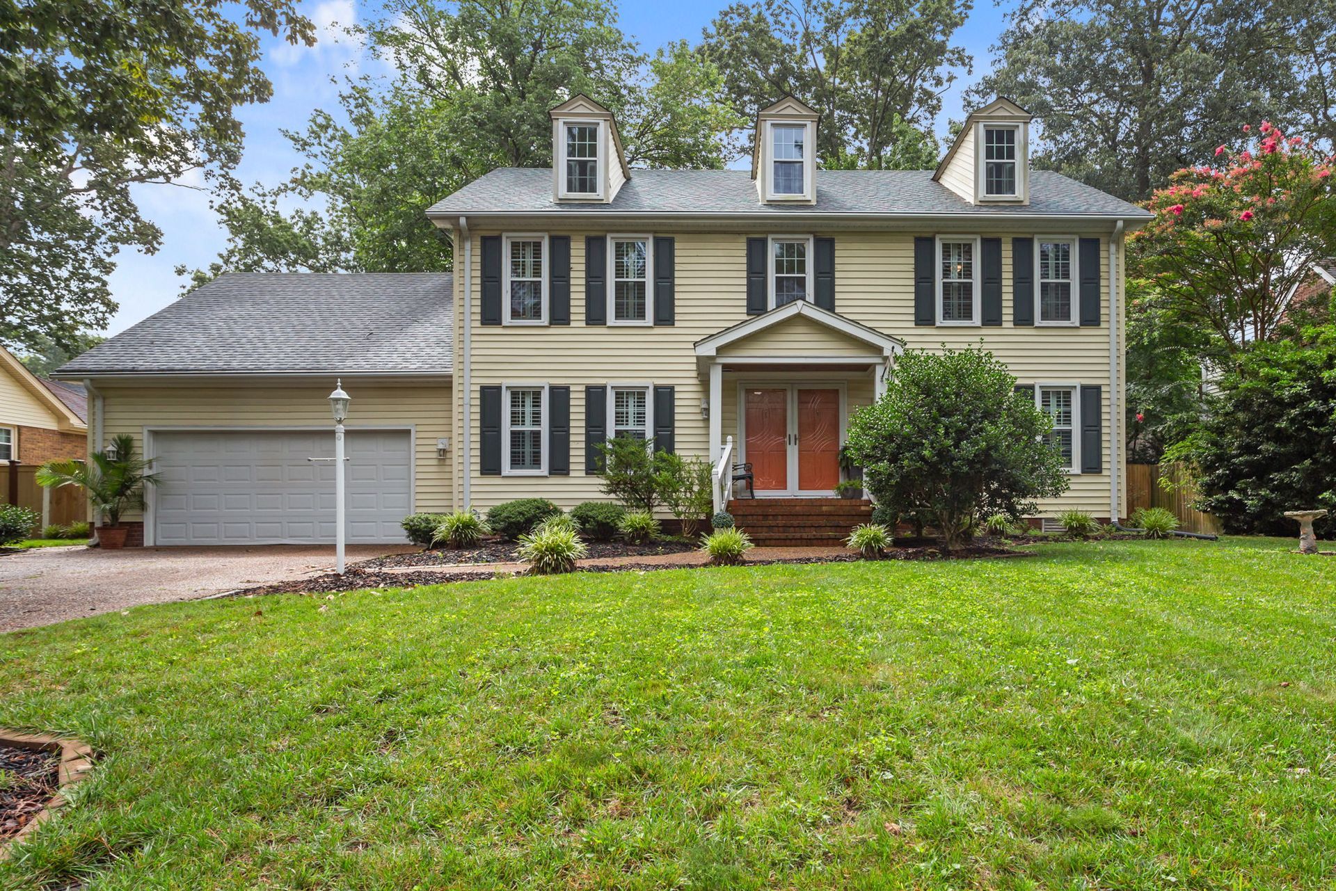 Two-story beige house with black shutters, attached garage, and a green lawn under trees