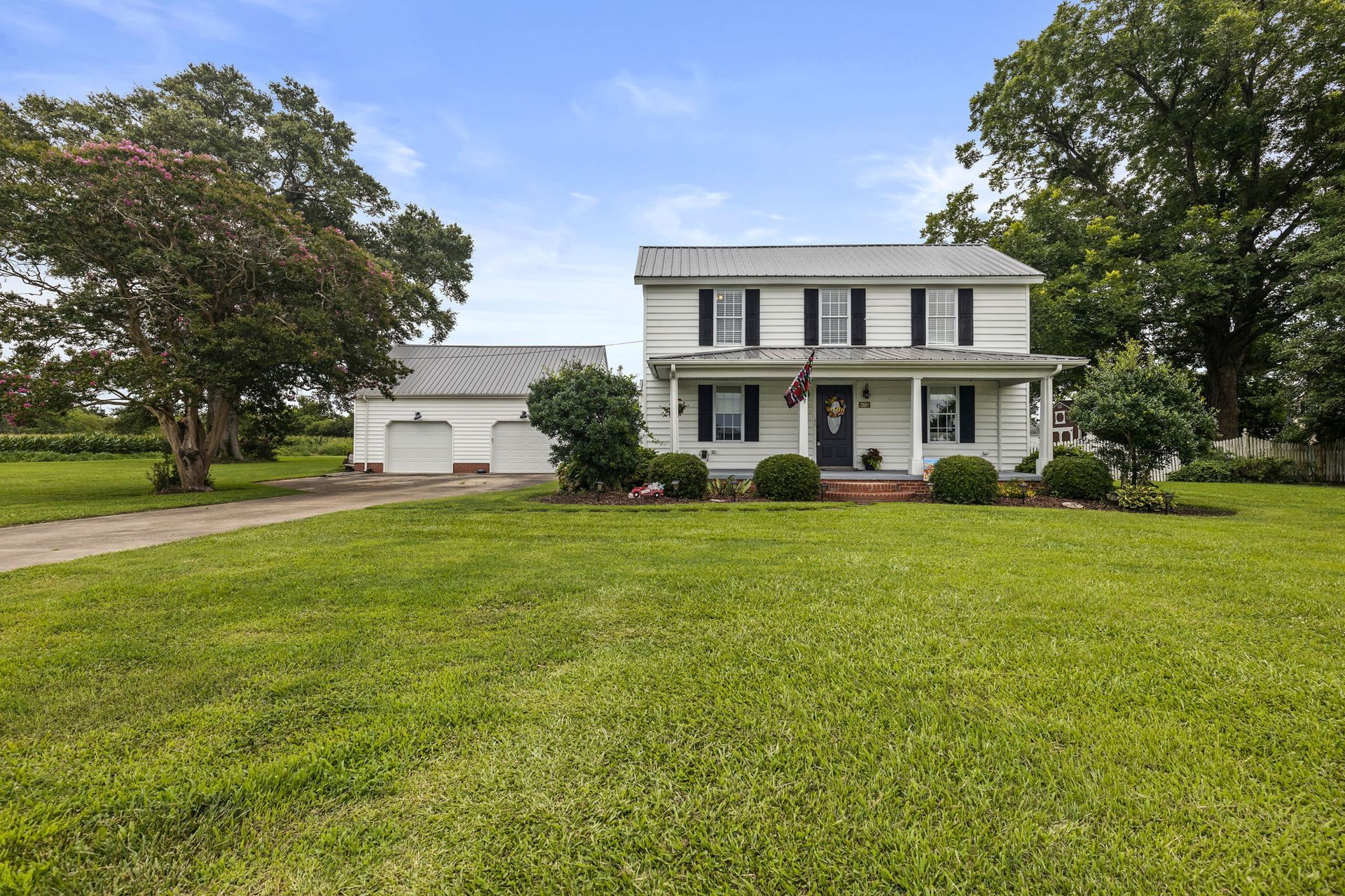 Two-story white farmhouse with attached garage, front porch, and large green lawn under a blue sky