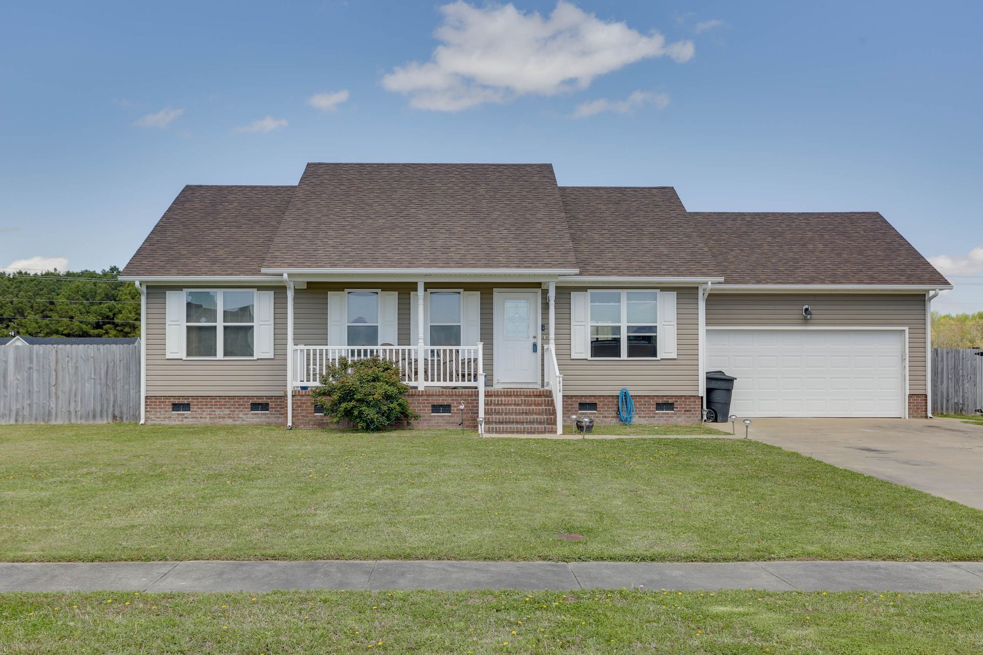 Single-story suburban house with a front porch, attached garage, and large front lawn under a blue sky