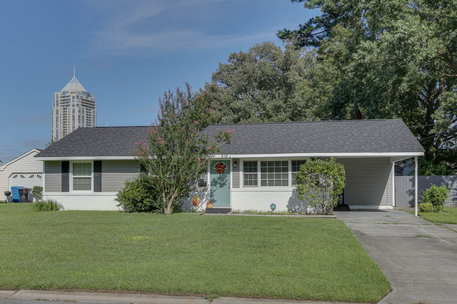 Single-story gray house with green lawn, driveway, and a tall building in the background