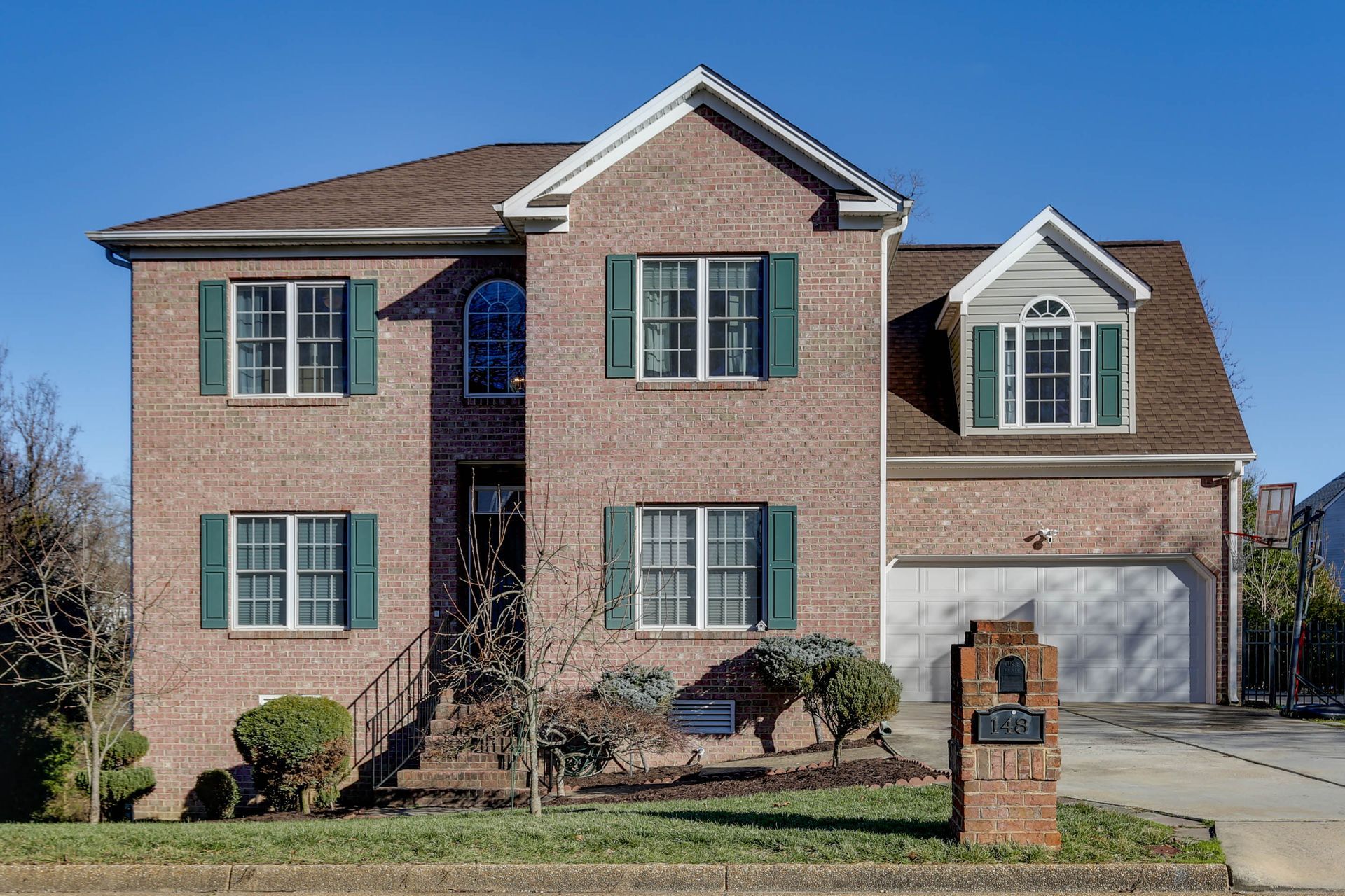 Two-story brick house with green shutters, front lawn, and attached garage under a clear blue sky