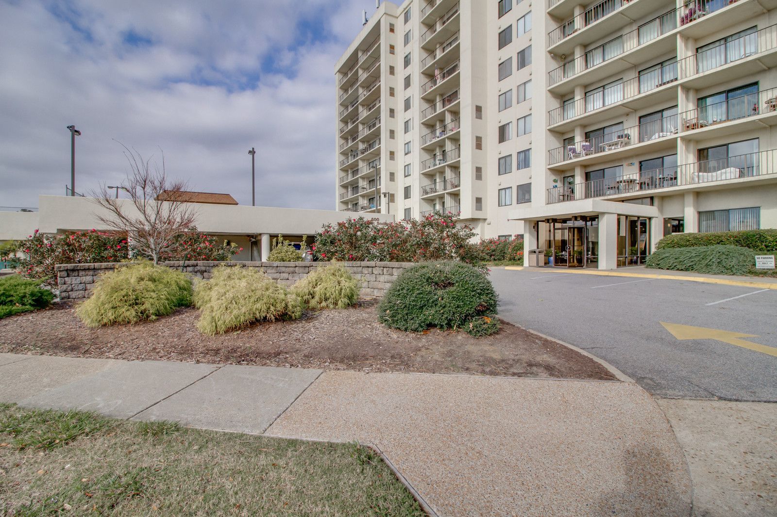 Apartment entrance with landscaped shrubs, sidewalk, and tall residential towers under a cloudy sky