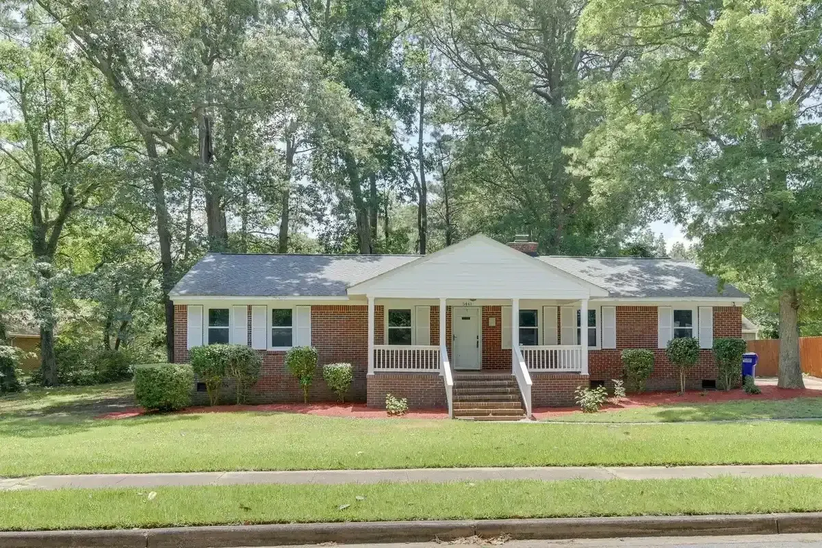 Single-story brick house with white trim, front porch, and large trees in a sunny yard.