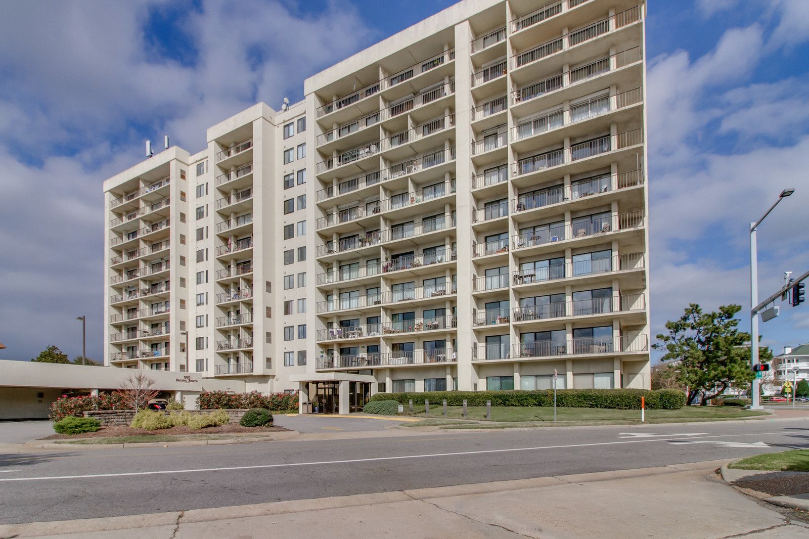 Tall beige apartment building with balconies beside a street under a partly cloudy sky
