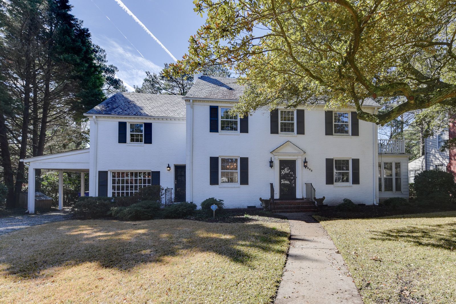 White two-story house with black shutters, front walkway, and large trees in a sunny yard