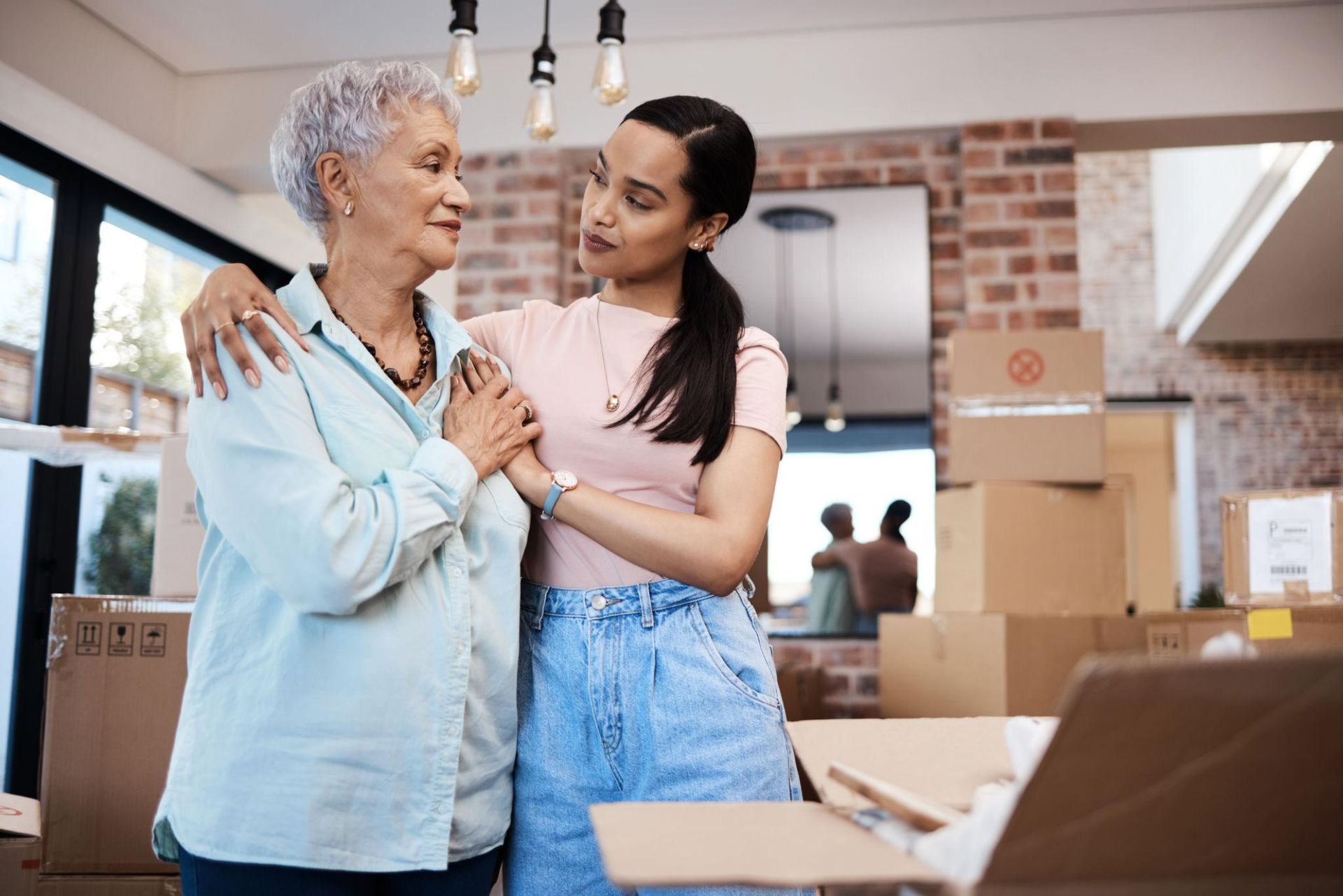 Two women smiling and embracing in a bright room with moving boxes and cardboard packages