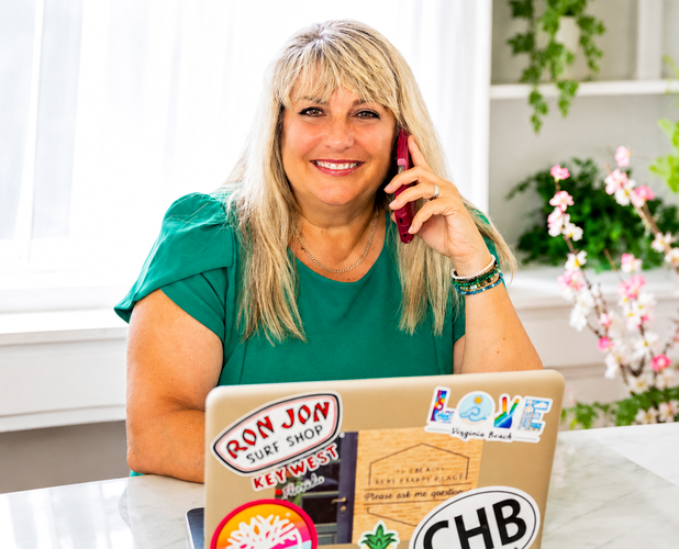 Woman in green shirt smiling at laptop with stickers, holding a phone in a bright office.