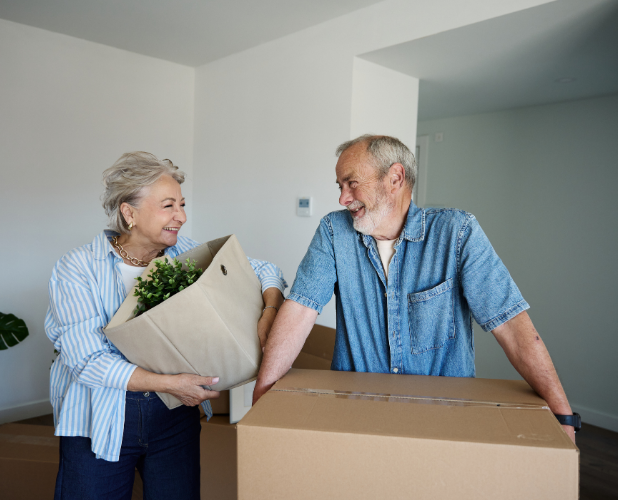 Smiling older couple packing boxes in a bright room, one holding a potted plant.