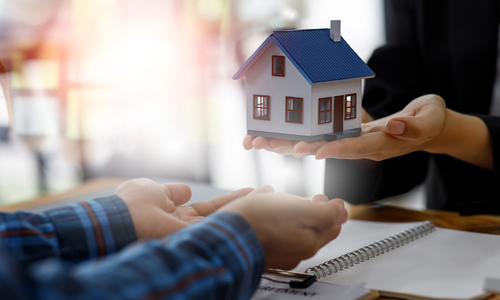 Hands exchanging a small house model over a desk with paperwork, suggesting a real estate transaction