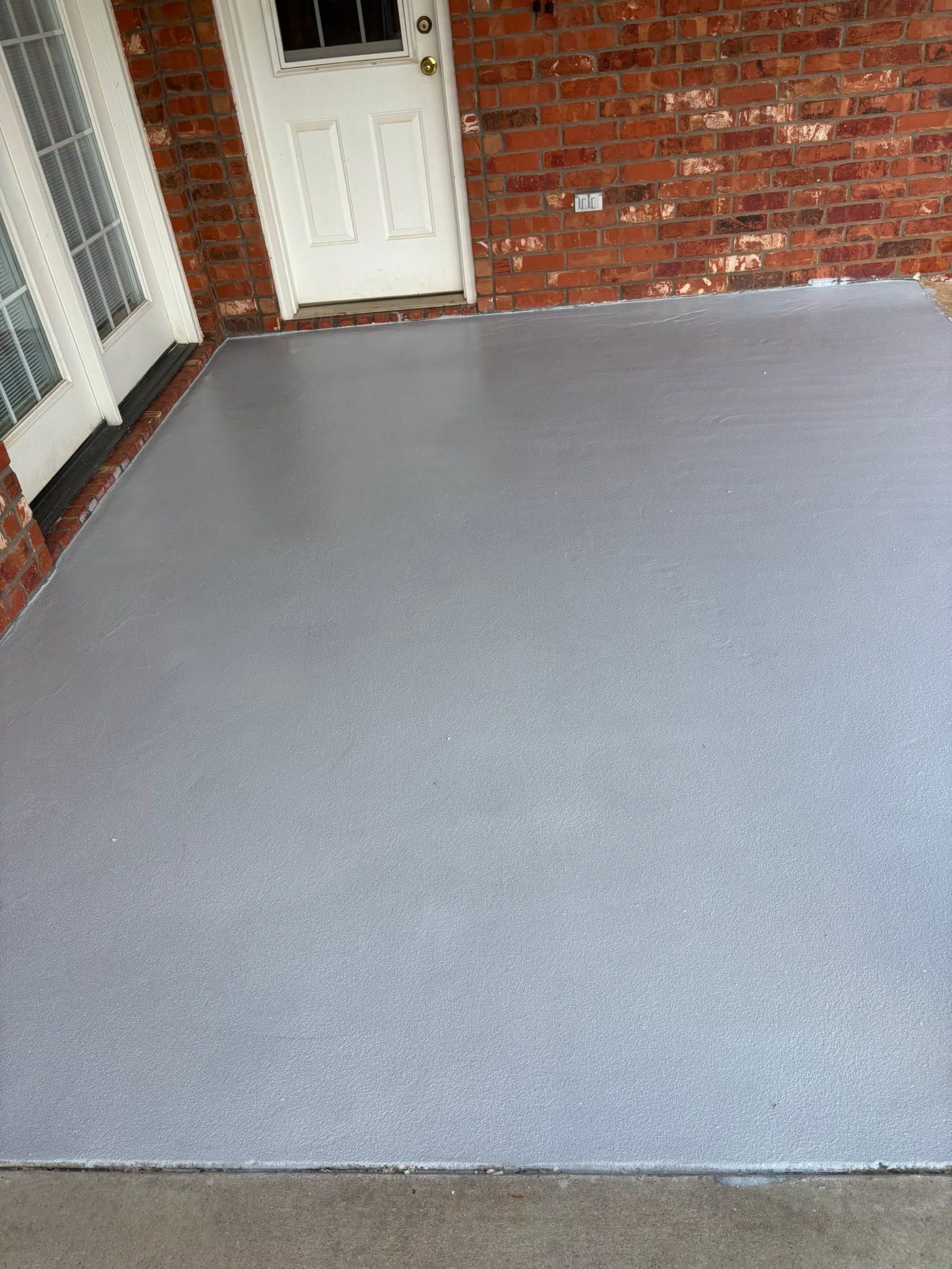Covered front porch with gray textured flooring, brick walls, and a white door.