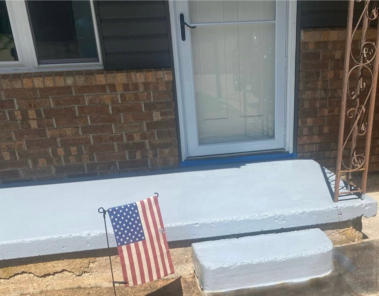 Front porch with white steps, blue-trimmed door, brick wall, and a small American flag in the ground.
