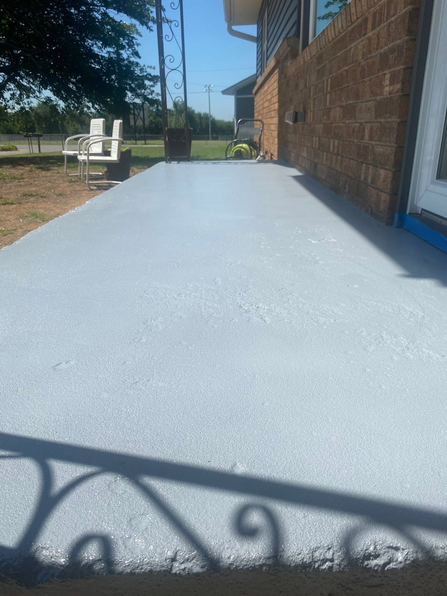 Freshly painted light blue concrete patio beside a brick house with yard furniture and a metal railing shadow