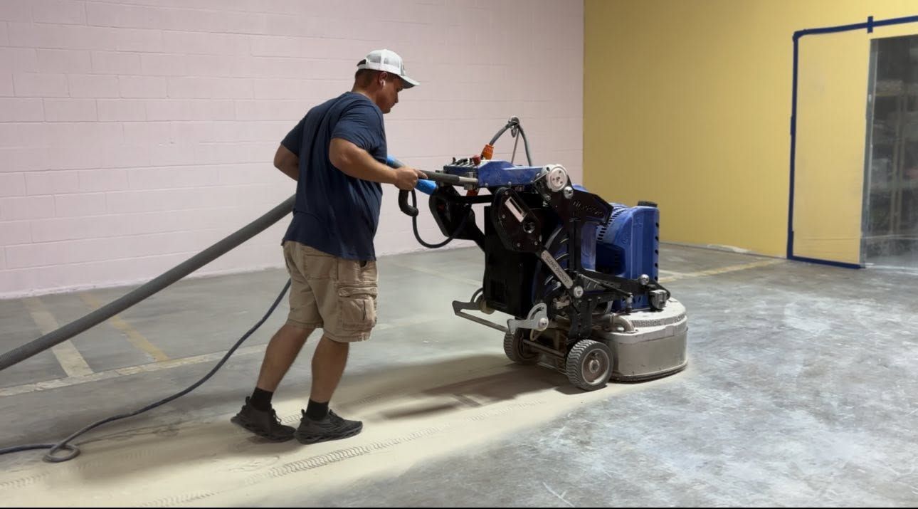 Worker operates a floor grinder in a dusty indoor room with a gray floor and yellow wall.