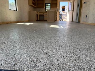 Empty unfinished room with speckled concrete floor, kitchen area, and open doorway to outside.