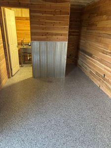 Wood-paneled room with gravel floor, open doorway, and a metal stove visible in the adjacent room