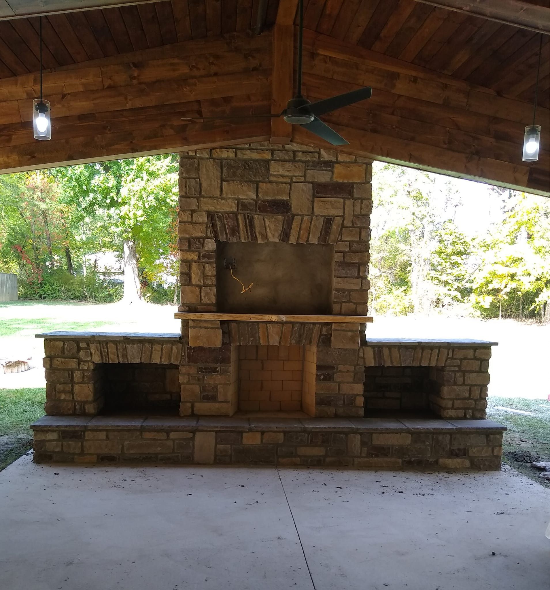 A stone fireplace under a wooden roof with a ceiling fan