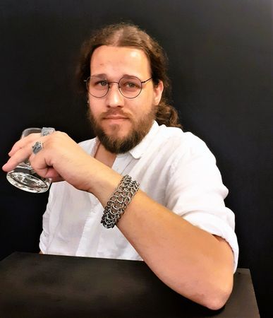 Persoon in een wit shirt met bril, zittend aan een tafel en een helder glas vasthoudend tegen een donkere achtergrond.