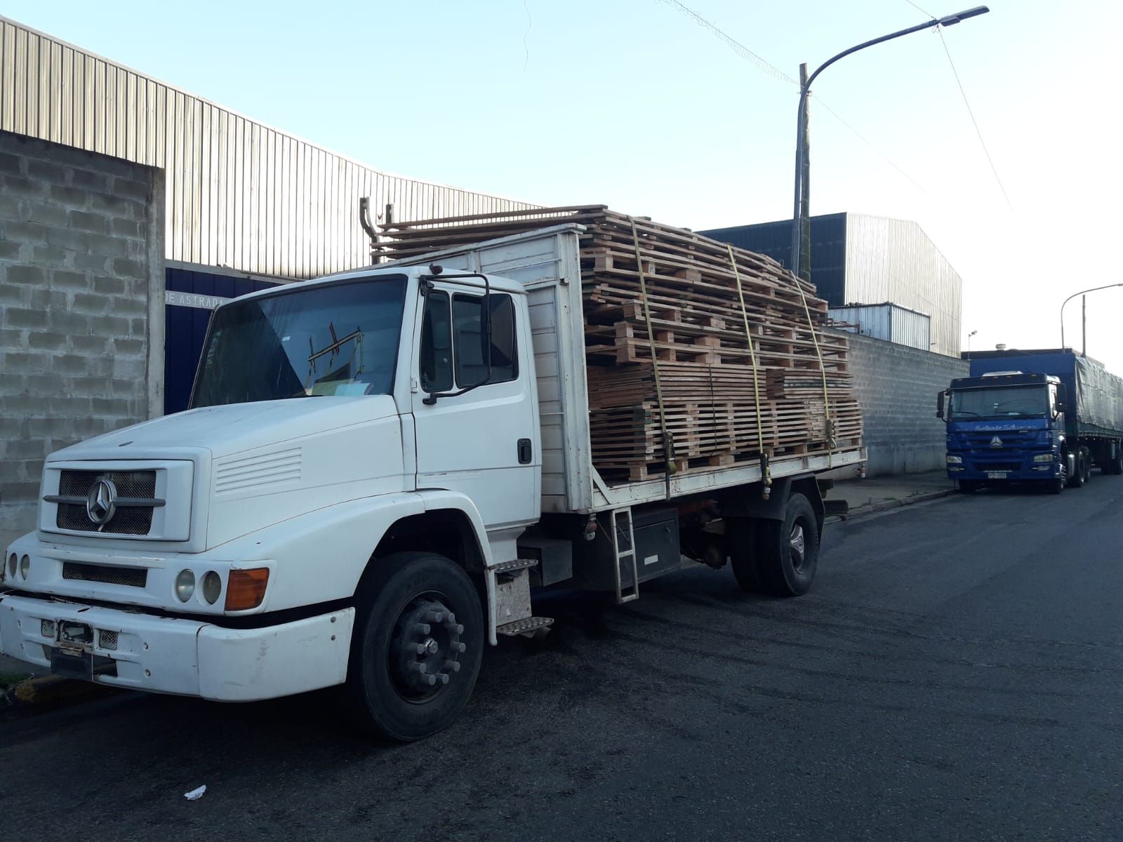 Un camión blanco cargado con una alta pila de palés de madera estaba estacionado en una calle junto a un camión azul.
