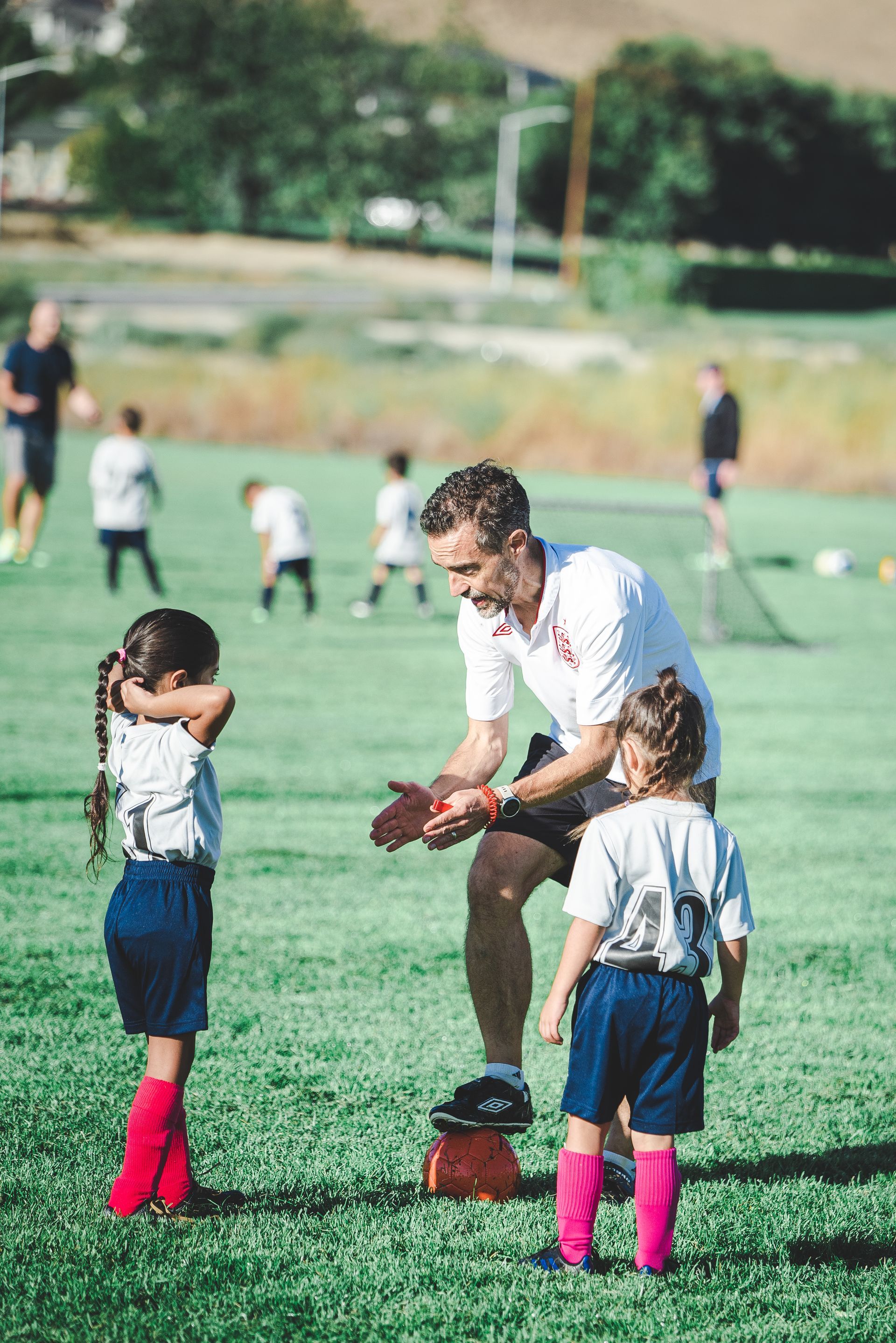 Coach instructing two children on a grassy field while others play in the background