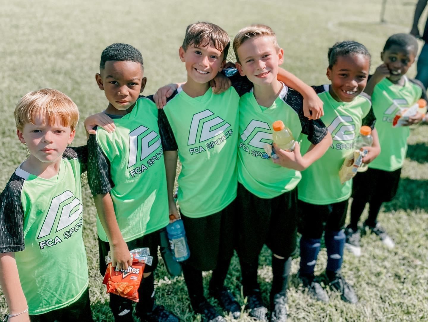 Five children in matching green jerseys stand on a grassy field, posing for a team photo.