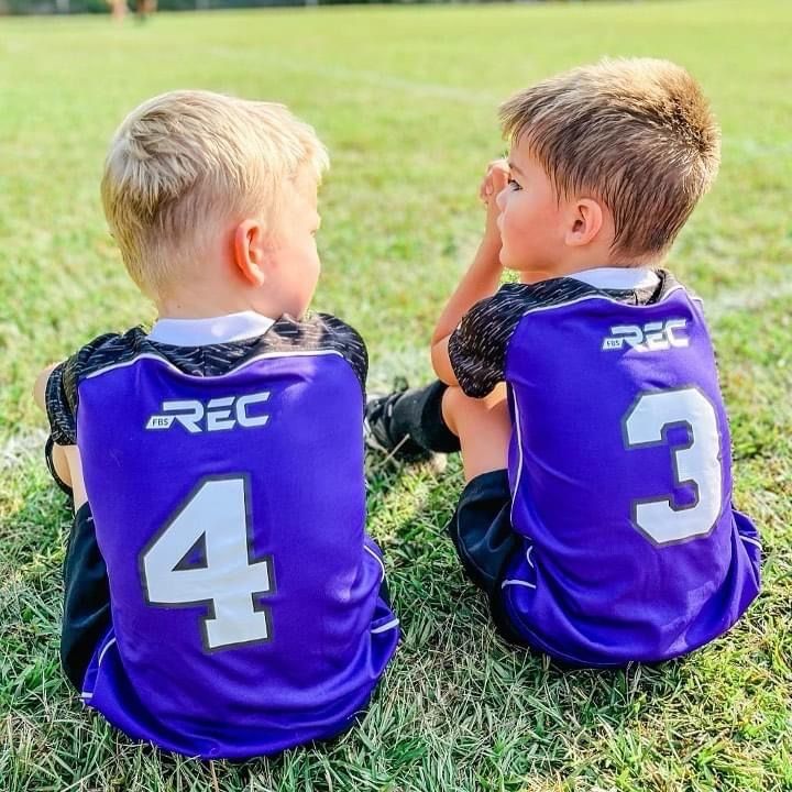Two children in purple REC jerseys sit on grass, seen from behind, with numbers 4 and 3 visible.