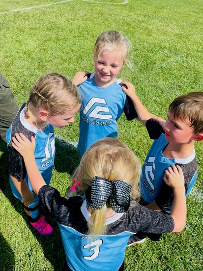 Four children in blue sports jerseys huddle together on a grassy field, smiling.