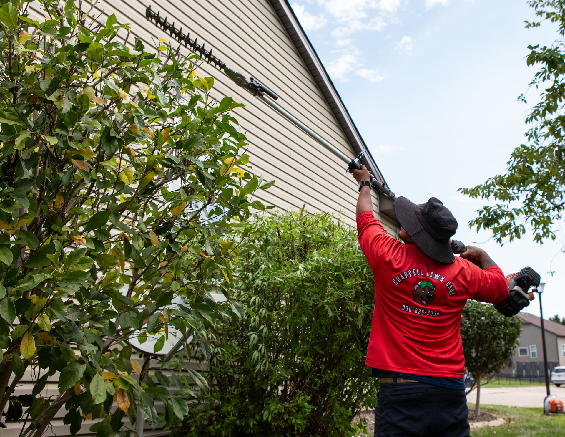 Worker in red jacket trimming tree branches beside a house with a pole saw
