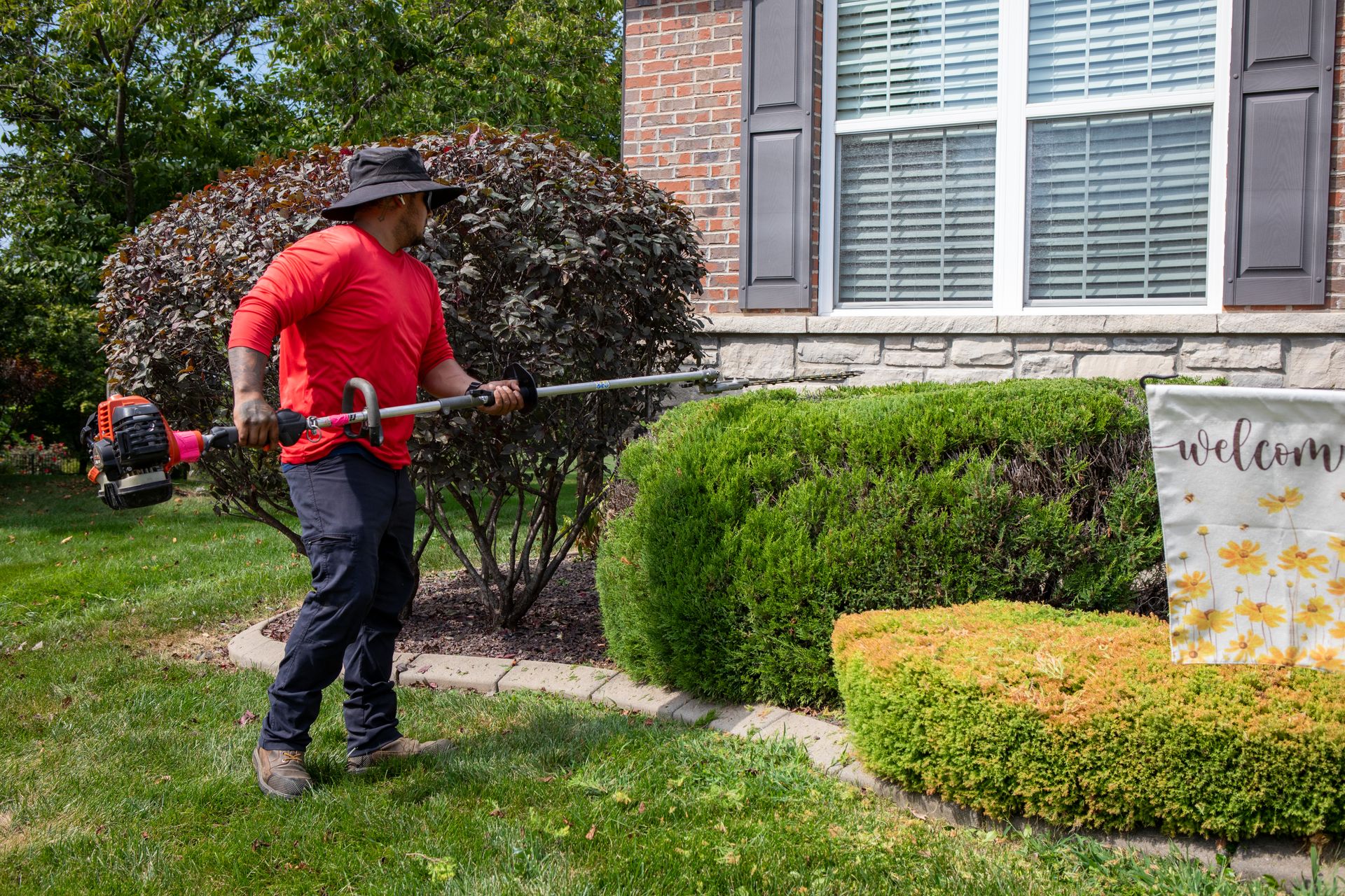 Person using a hedge trimmer to shape bushes beside a house and welcome sign