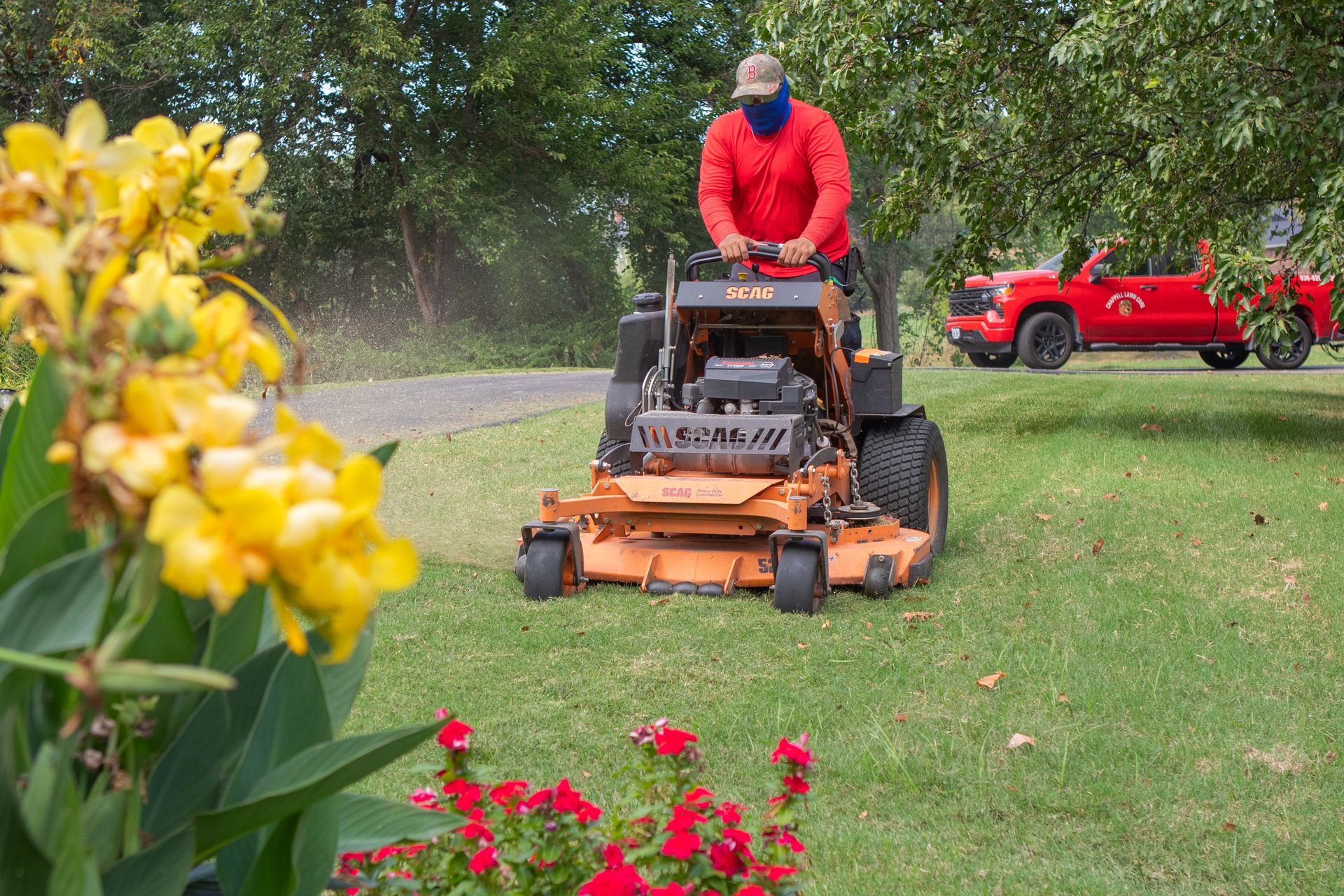 Worker riding orange mower on a grassy lawn, with flowers in foreground and red truck in background