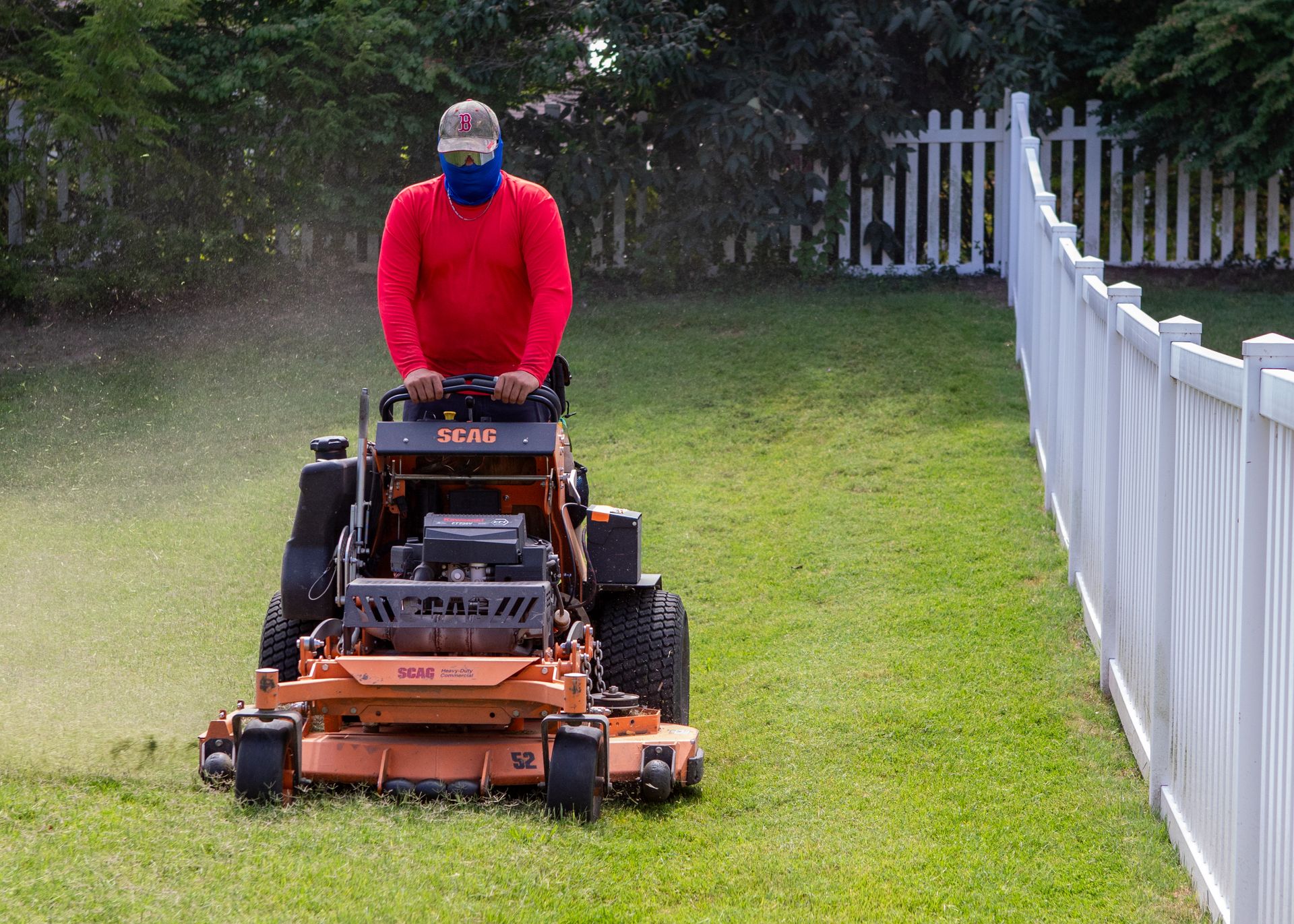 Person mowing a grassy lawn with an orange riding mower beside a white fence