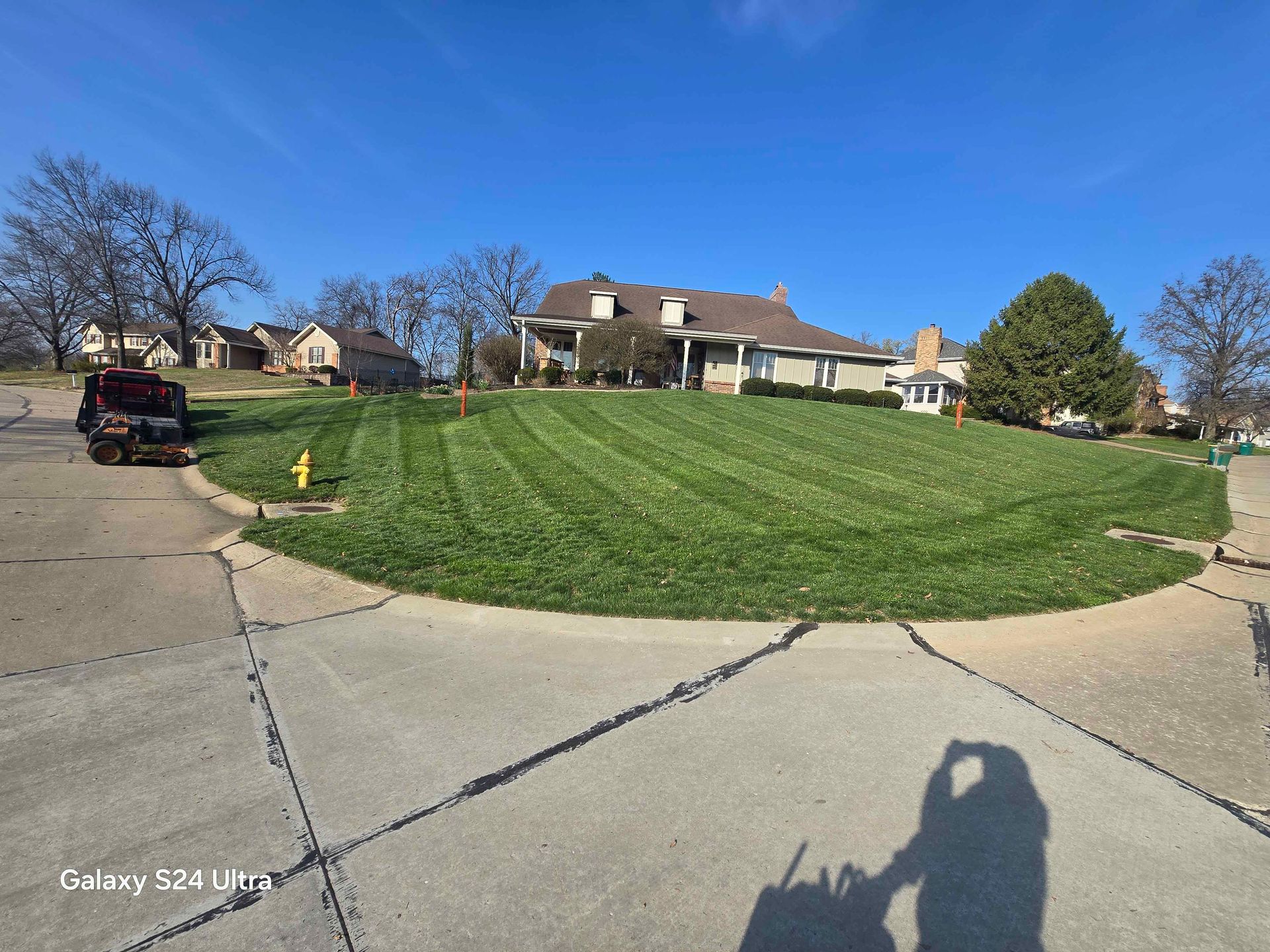 Suburban street with a grassy corner lot, houses, parked SUV, and a long shadow on the sidewalk under a blue sky