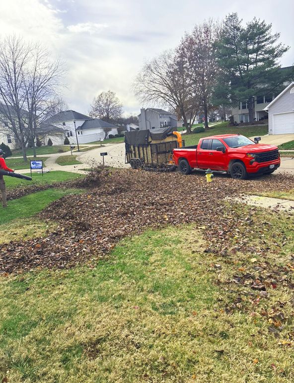 A red truck parked near a large pile of leaves as a worker blows leaves. Residential setting.