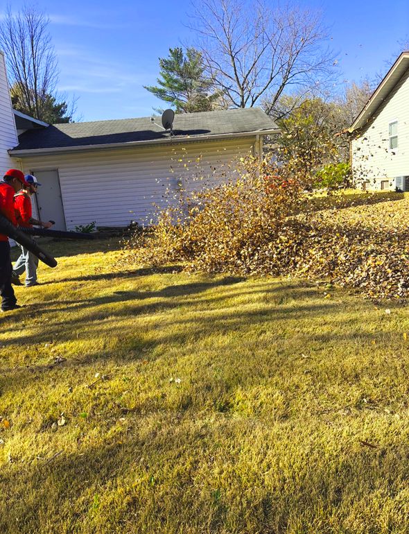 Two people blowing leaves on a lawn; a white garage and house are visible in the background.