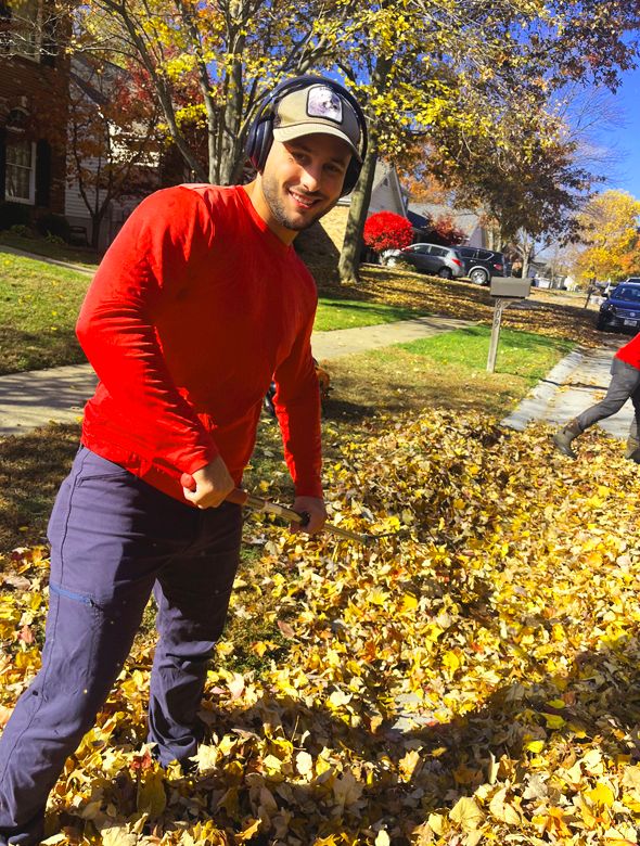 Man in red shirt and blue pants raking leaves on a sunny fall day.