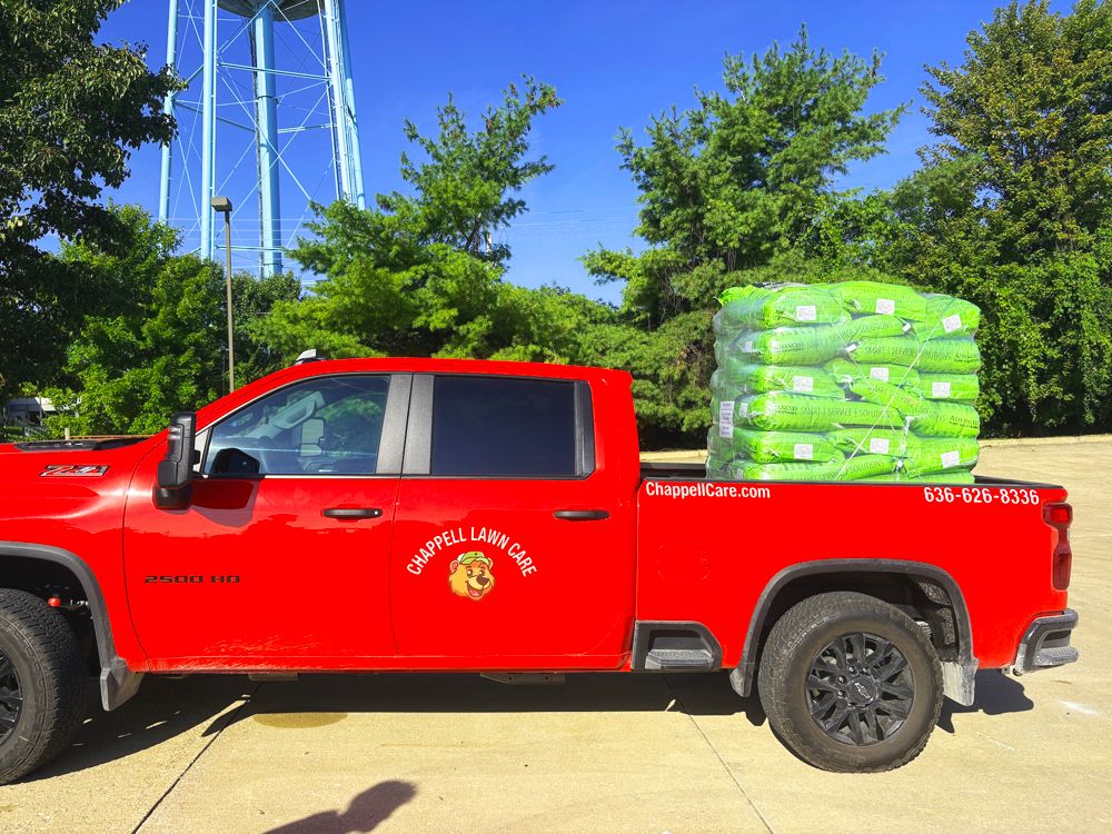 Red Chapel Lawn Care truck with a pallet of green bags in the bed, parked near a water tower.